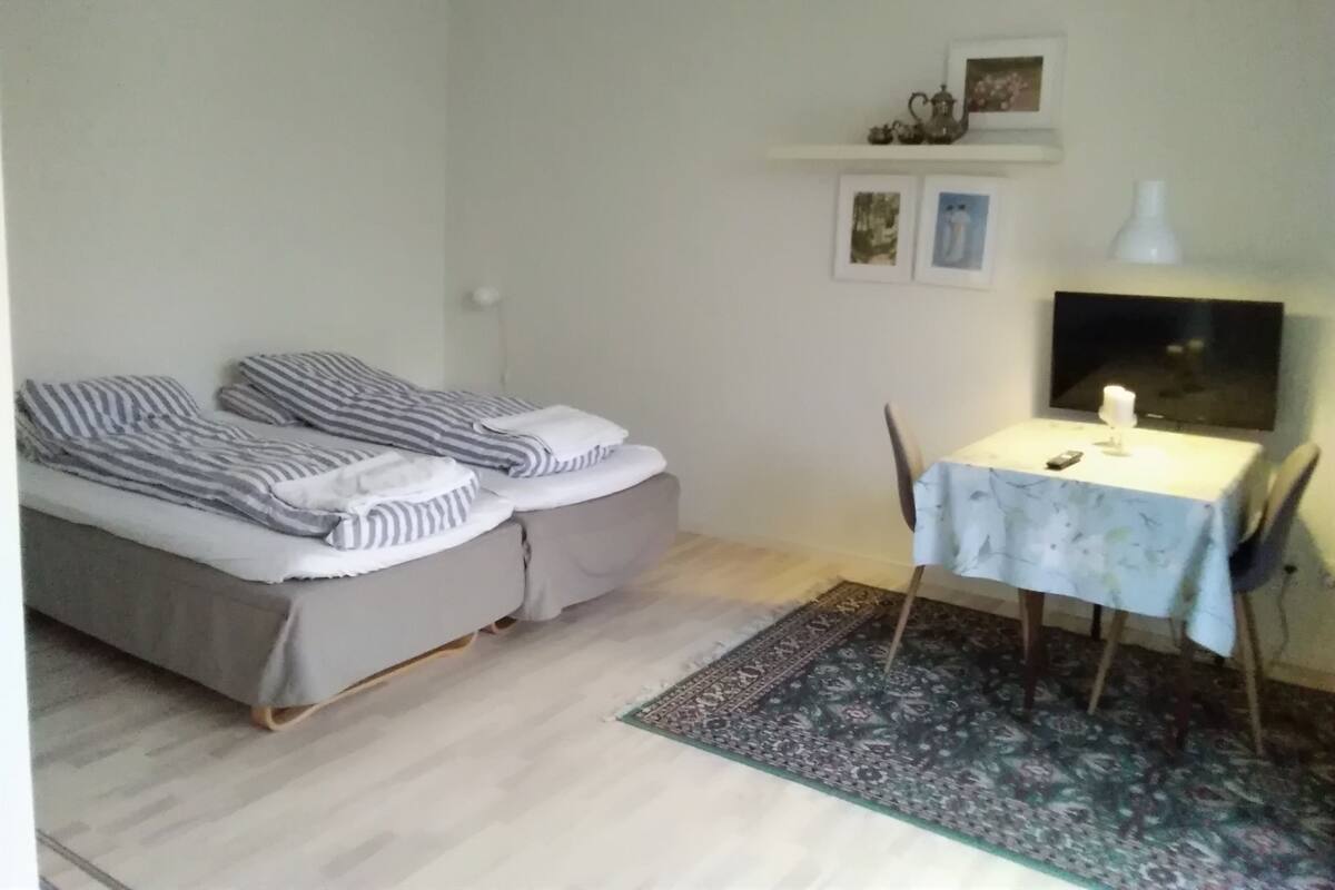 Una habitación pequeña y ordenada con una cama, una mesa con una lámpara y una silla. Hay cuadros enmarcados en la pared y una pequeña estantería sobre la cama.