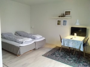 Una habitación pequeña y ordenada con una cama, una mesa con una lámpara y una silla. Hay cuadros enmarcados en la pared y una pequeña estantería sobre la cama.