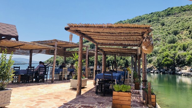 Een buiten eetgedeelte met houten tafels en stoelen, overdekt met rieten parasols, met op de achtergrond een meer en heuvels.