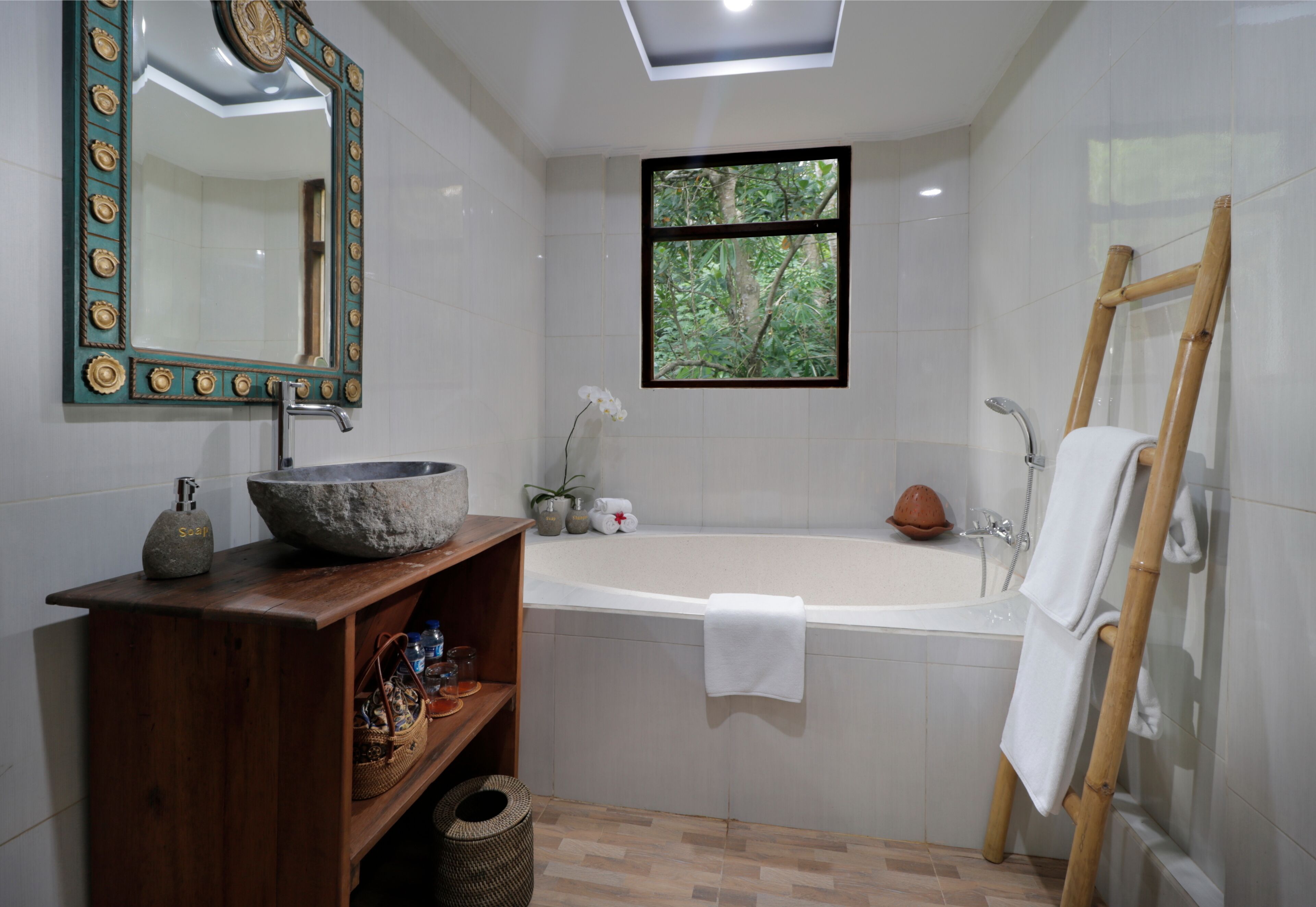A bathroom with a bathtub, a wooden vanity, a large mirror, a window with a view of trees, and a bamboo ladder.