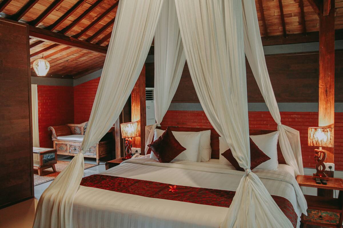 Una cama con dosel y cortinas, ropa de cama blanca con detalles rojos, cabecera de madera y pared de ladrillo rojo.