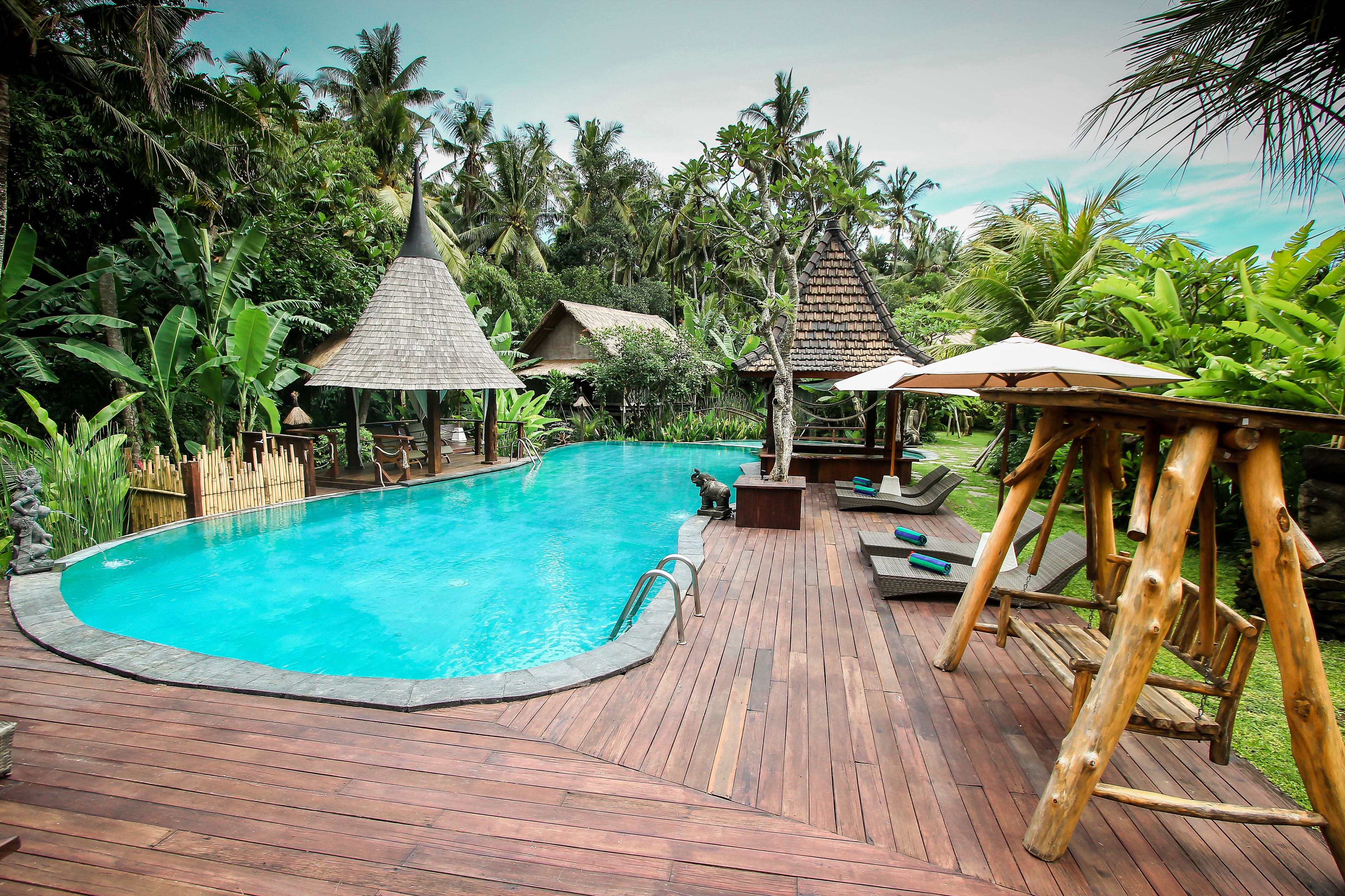 A wooden deck surrounding a swimming pool with thatched-roof structures and lush greenery.