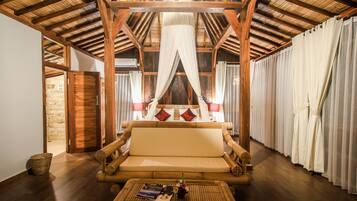 Una habitación con techo de madera, una cama con cortinas blancas, un sofá y una mesa de mimbre con un libro y flores.