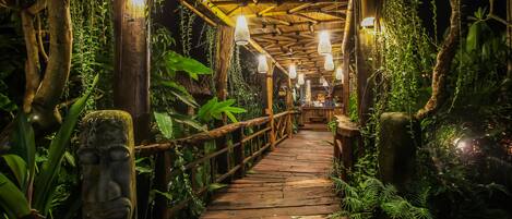 A wooden pathway with hanging lights, surrounded by lush greenery and a stone face sculpture.