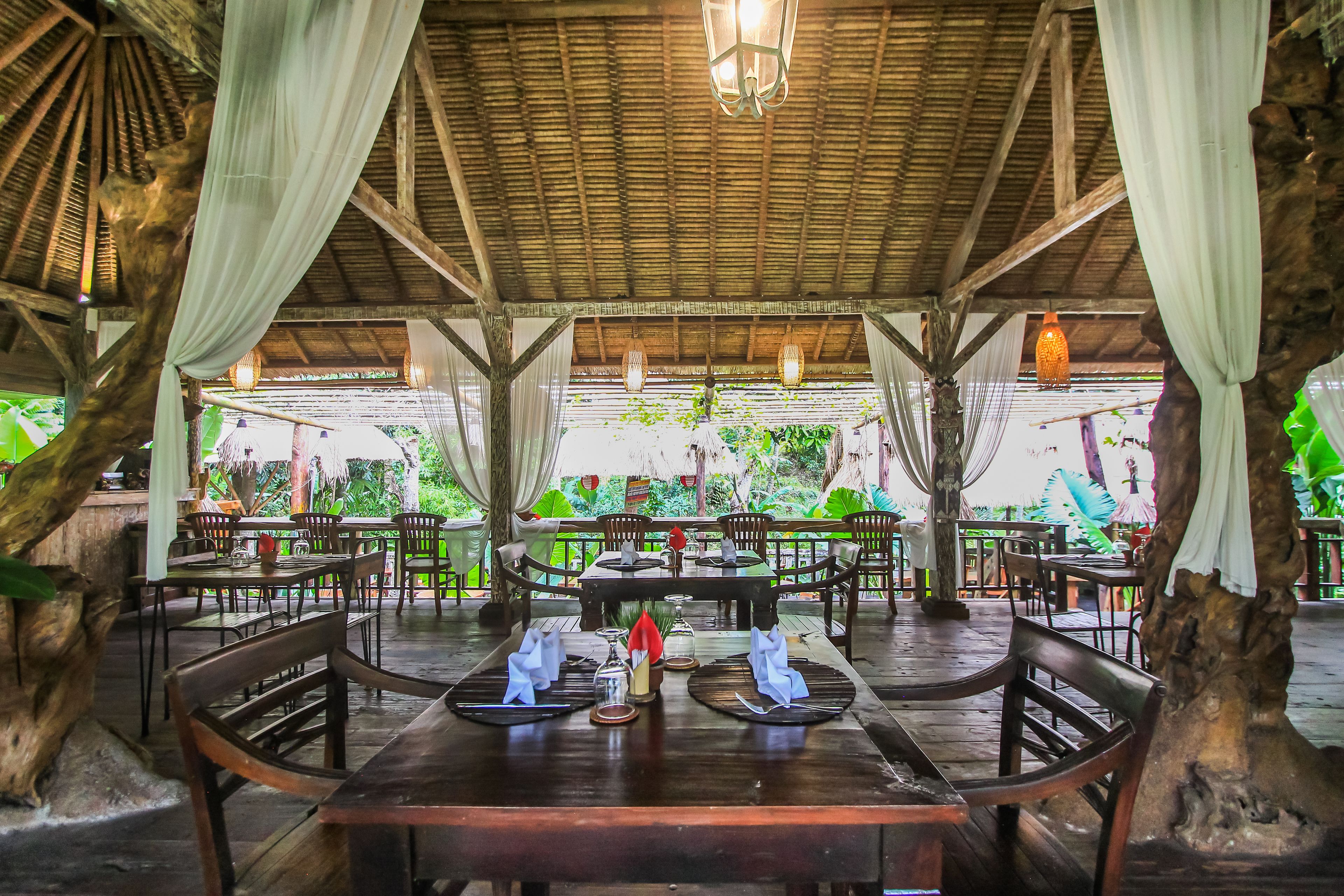 An outdoor restaurant with wooden tables and chairs, draped with white curtains, and surrounded by lush greenery.