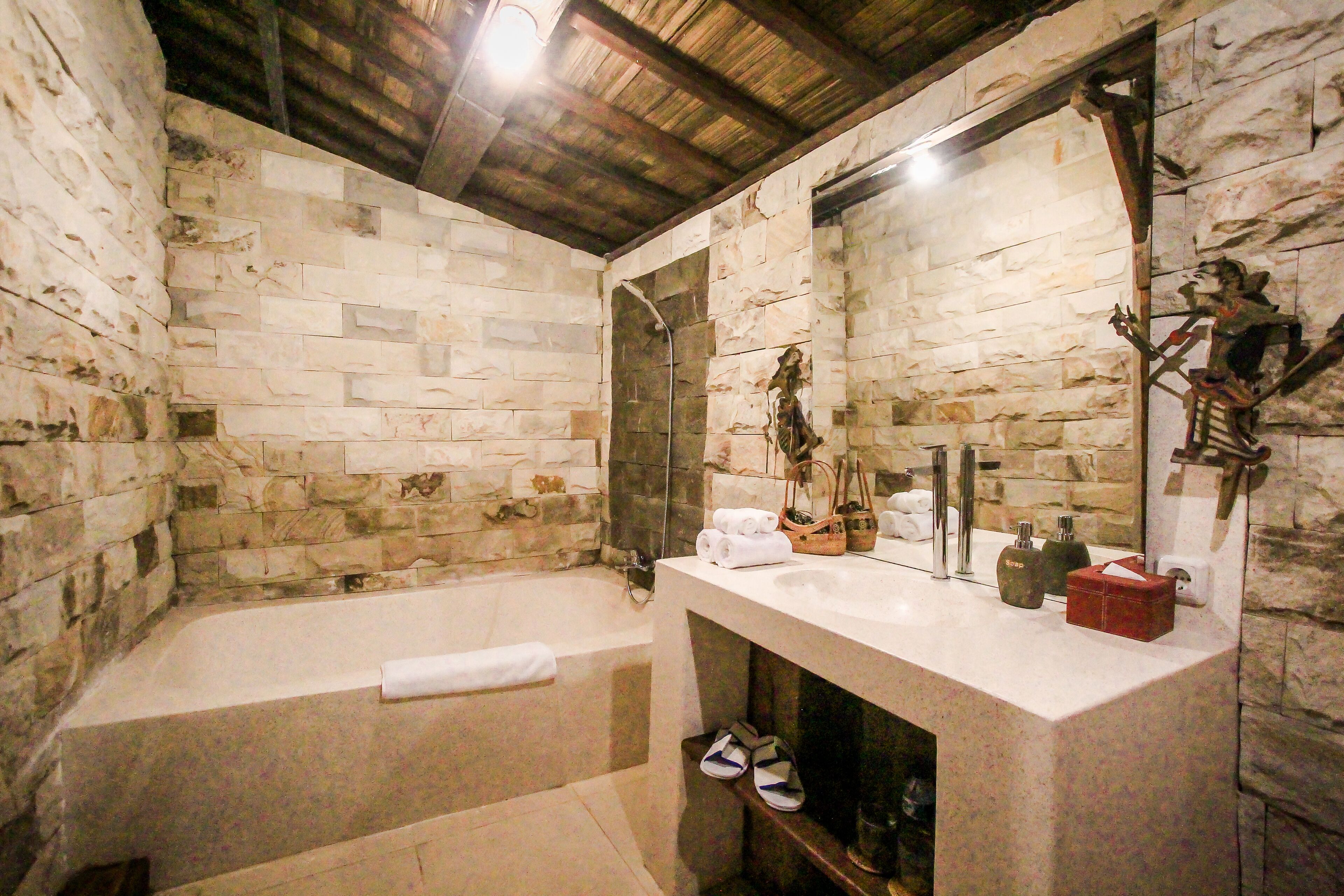 A stone-walled bathroom with a bathtub, a large mirror, and a wooden shelf with toiletries.