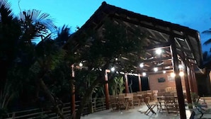 Un espace extérieur abrité, aménagé avec du mobilier en bois et doté d’une scène, le tout entouré d’arbres.