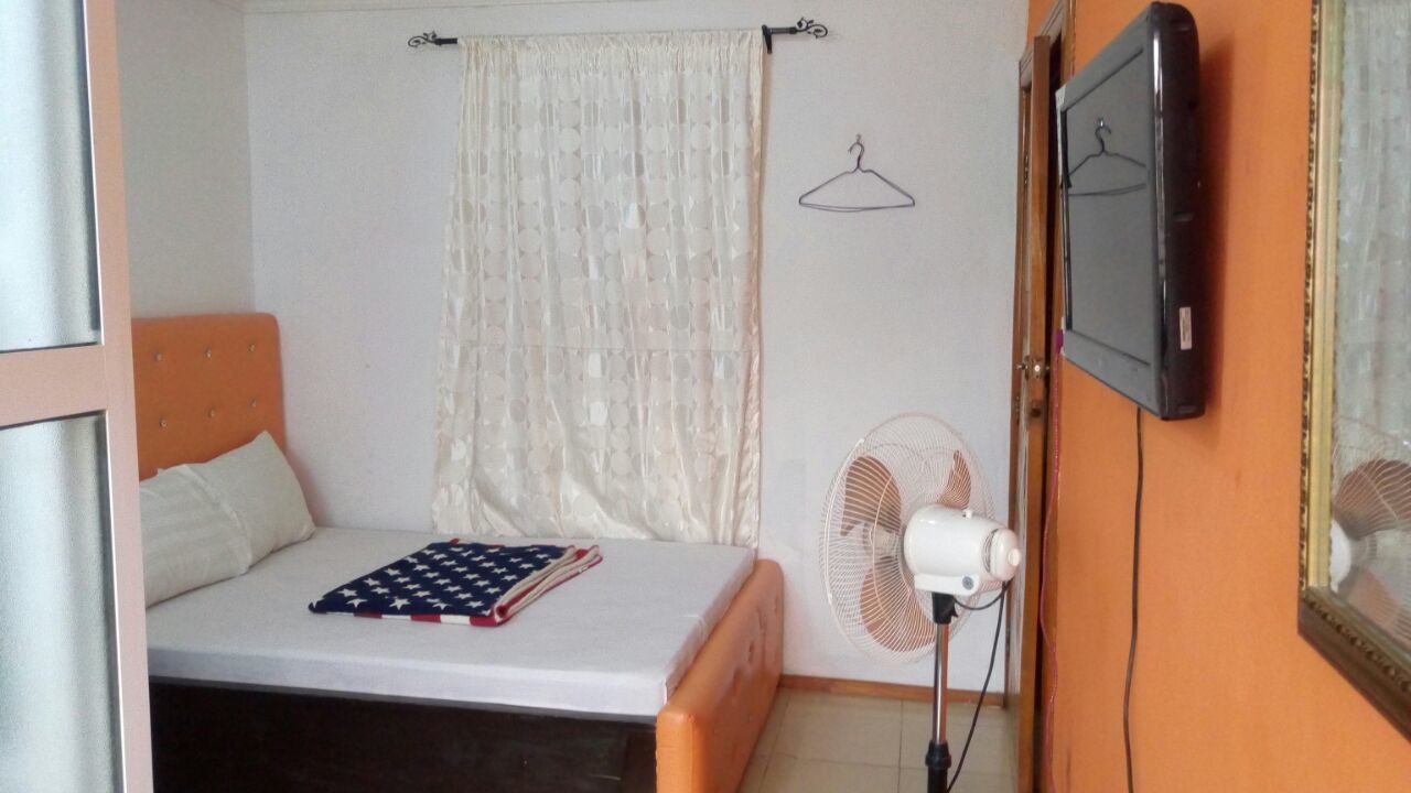 Quarto pequeno com uma cama de solteiro, colcha branca, um ventilador e uma televisão fixada na parede.