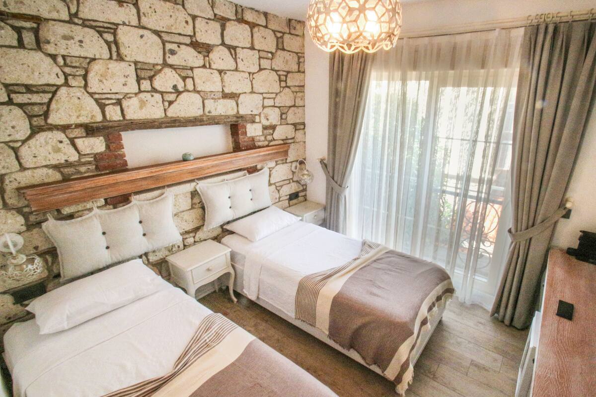 A bedroom with two beds, stone wall, wooden headboard, bedside tables, and a hanging light fixture.