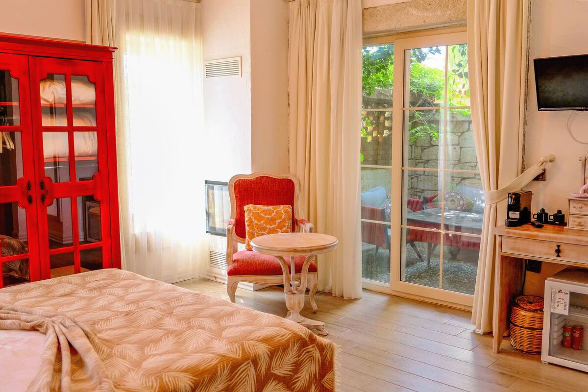 A hotel room with a bed, a red wardrobe, a small round table with a chair, a television, a mirror, and a window with a view of greenery.