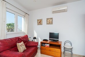A room with a red leather sofa, a wooden TV stand with a flat-screen TV, a metal chair, and an air conditioning unit on the wall.