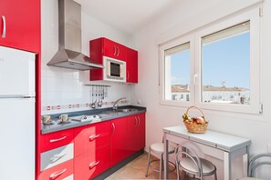 A compact kitchen with red cabinets, a white refrigerator, a microwave, and a window with a view of buildings.