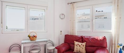 A room with a red leather sofa, a small table with a basket, two metal chairs, and a window with curtains.