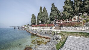 A coastal walkway with a wooden deck, clear water, stone walls, and tall trees. - Modern Apartment "Gennaker" On Lakeside with Terrace and View (Malcesine)
