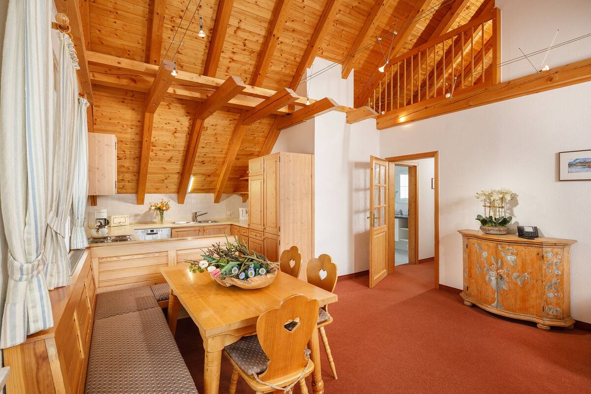 Un interior de madera con una mesa de comedor, sillas y un banco. Se observa un aparador con adornos florales y una puerta que da a otra habitación.