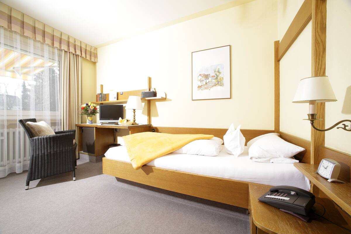 Habitación de hotel con cama, escritorio con lámpara, silla, mesita de noche con teléfono y ventana con cortinas.