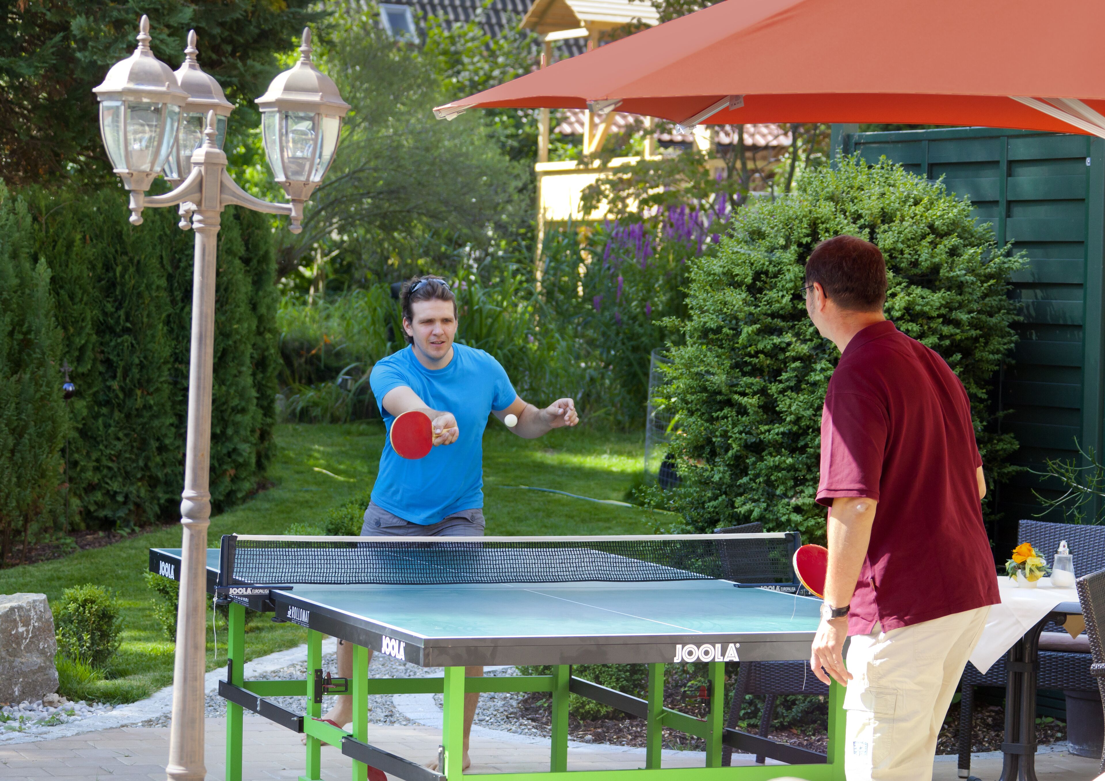 Una mesa de ping pong al aire libre con dos jugadores jugando, rodeada de vegetación y un pabellón.