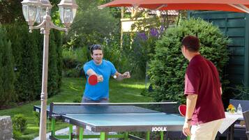 Una mesa de ping pong al aire libre con dos jugadores jugando, rodeada de vegetación y un pabellón.