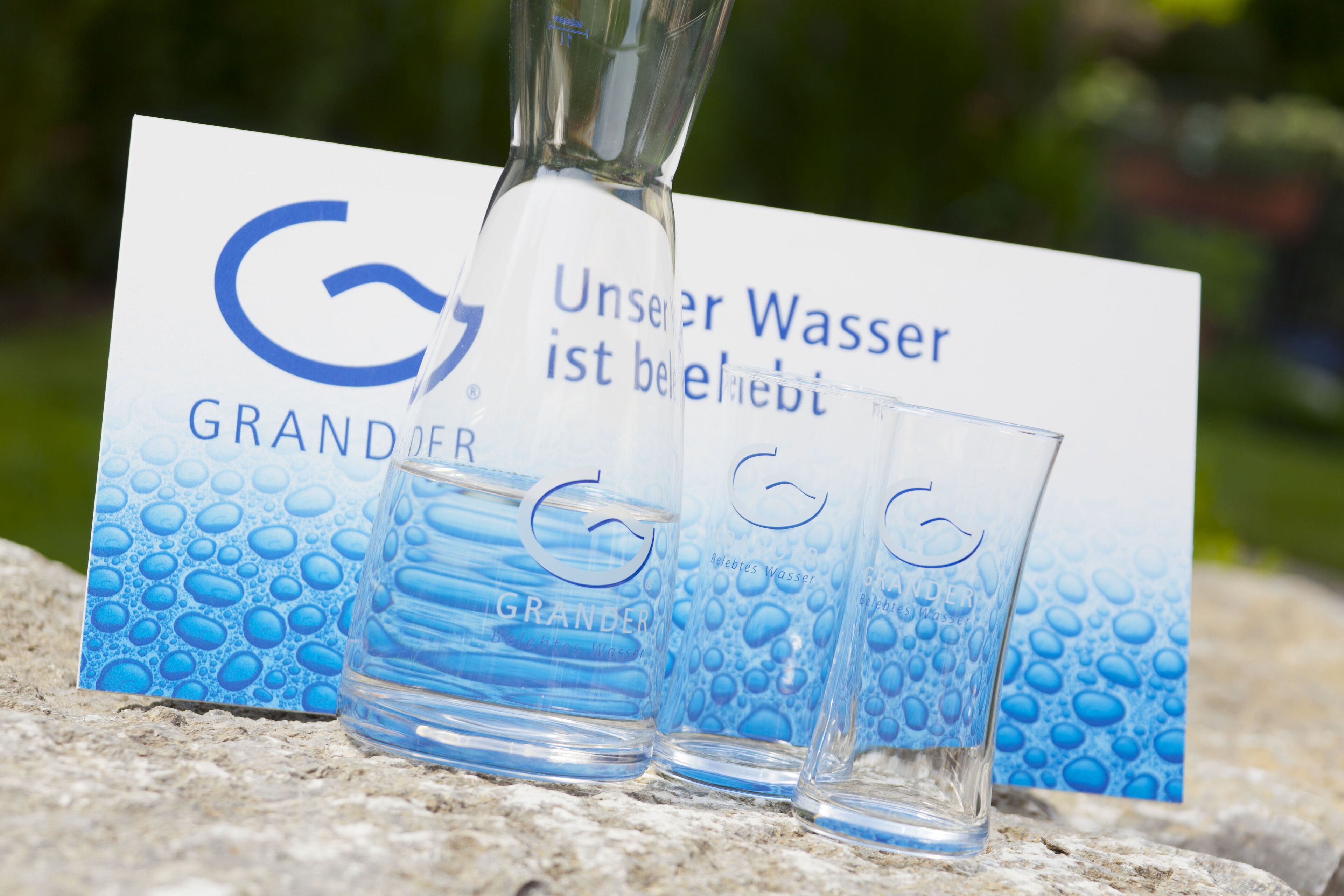 A glass bottle and two glasses with a water bottle logo, placed on a stone surface with a water bottle advertisement in the background.