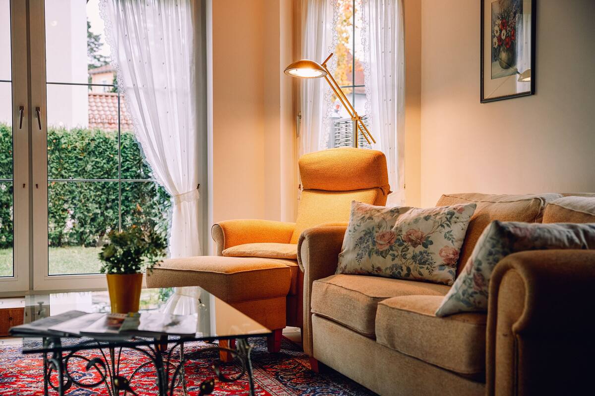 Un salón acogedor con un sofá, una silla, una mesa y una lámpara de pie. Se ve una ventana con cortinas y vistas al exterior con vegetación.