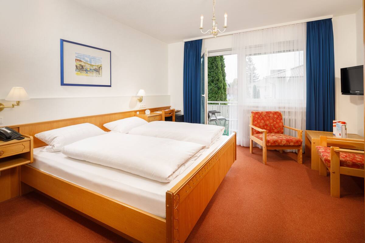 Habitación de hotel con cama de madera, dos mesitas de noche, una silla, un escritorio, un televisor y una ventana con cortinas.