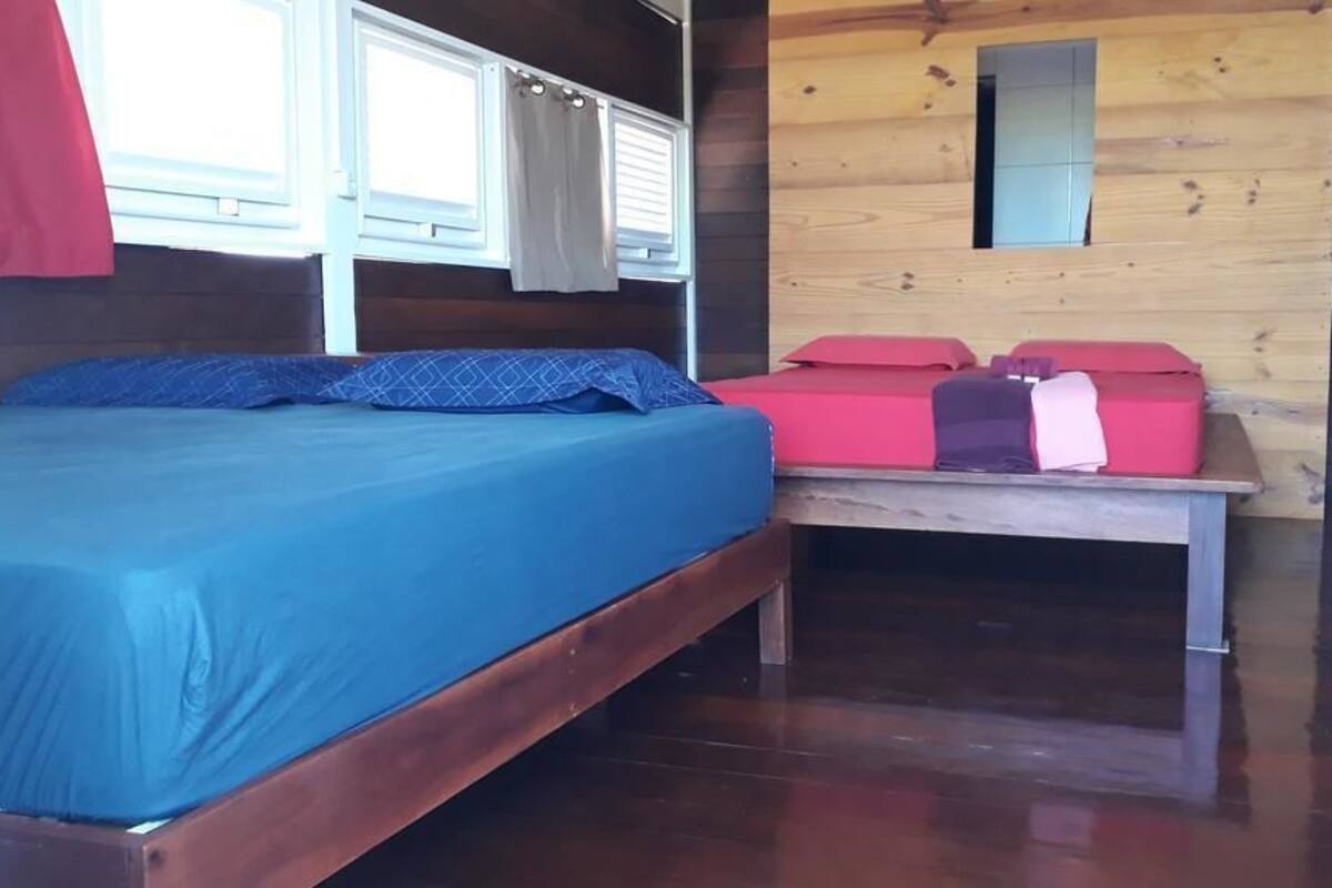 Une pièce avec un plancher en bois, un lit recouvert d’une courtepointe bleue, une petite table agrémentée de coussins rouges et roses, et une fenêtre avec des rideaux.