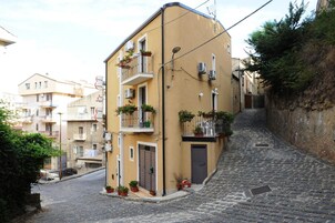 Una estrecha calle empedrada bordeada por edificios de varios pisos con balcones, algunos con plantas en macetas.