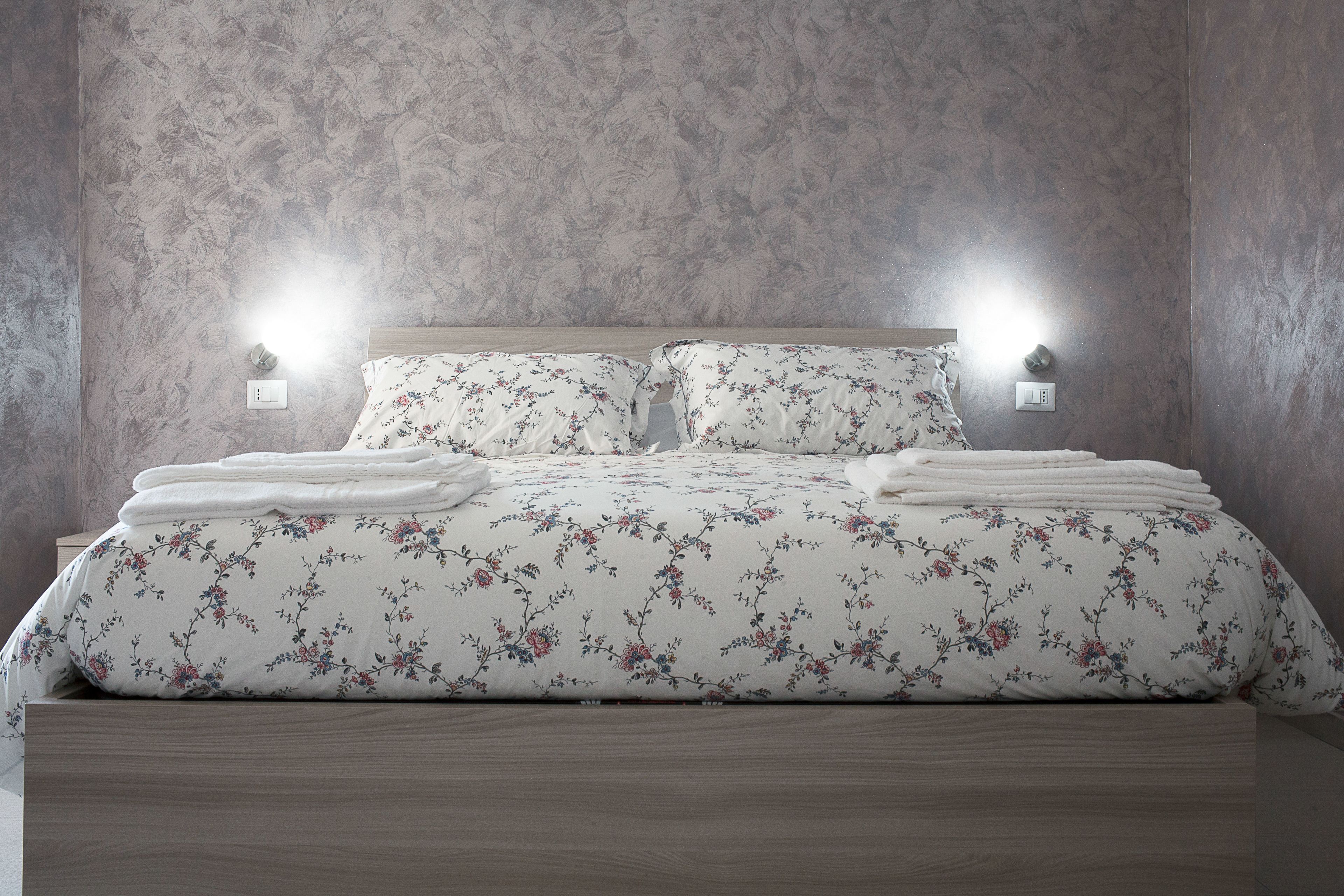 Una cama con ropa de cama floral, dos luces fijadas a la pared y un cabecero de madera.