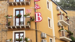 Um edifício amarelo com varandas decoradas com plantas em vasos, uma placa com a inscrição 'B&B la Regina di Saba' e uma rua de paralelepípedos.