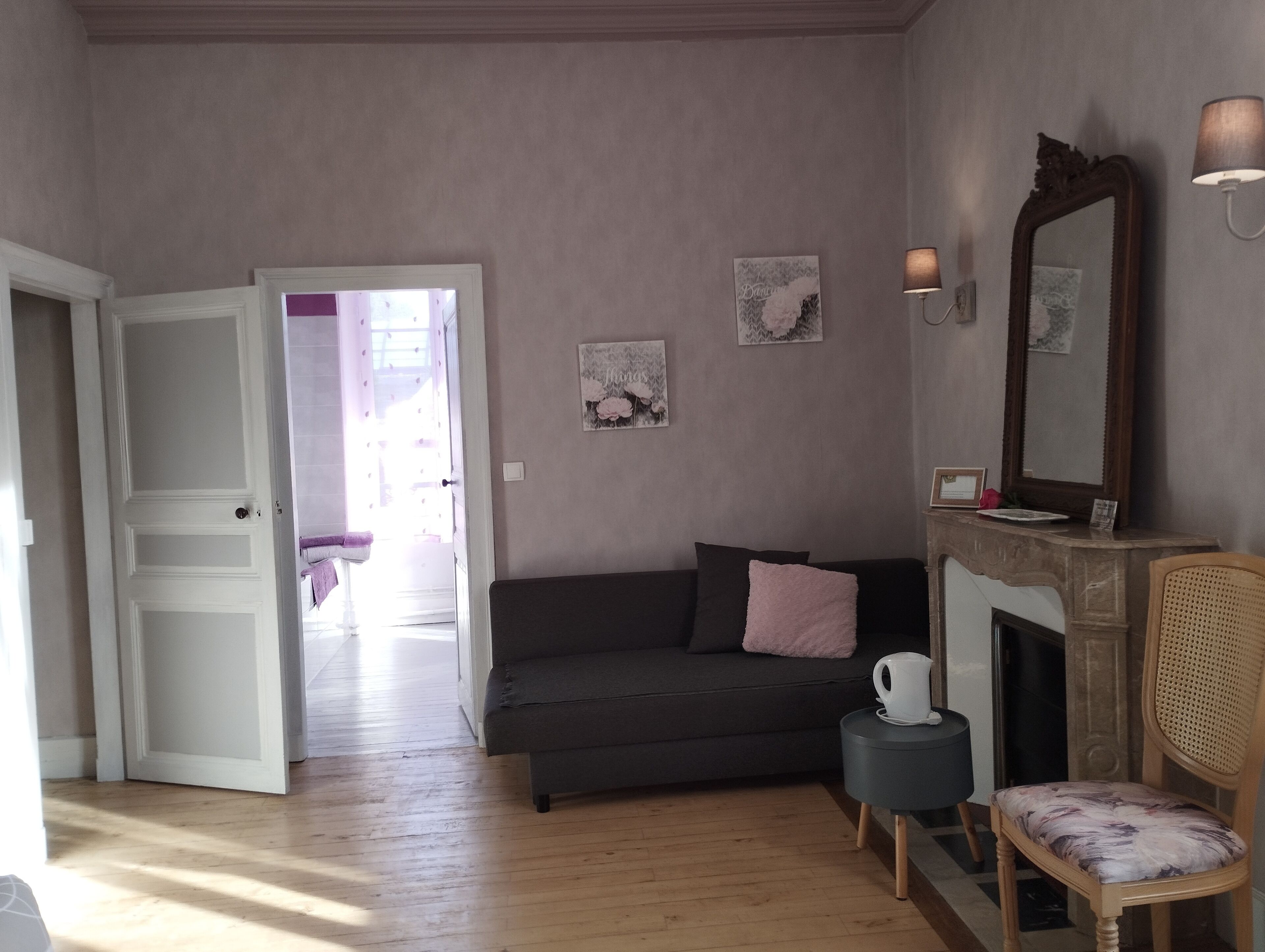 Ein Wohnzimmer mit einem dunkelgrauen Sofa, einem kleinen runden Tisch mit einem weißen Gegenstand, einem Holzstuhl und einem großen Spiegel an der Wand.