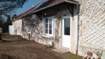 A rustic stone building with a sloped roof, a wooden door, and a small garden area with a dog.
