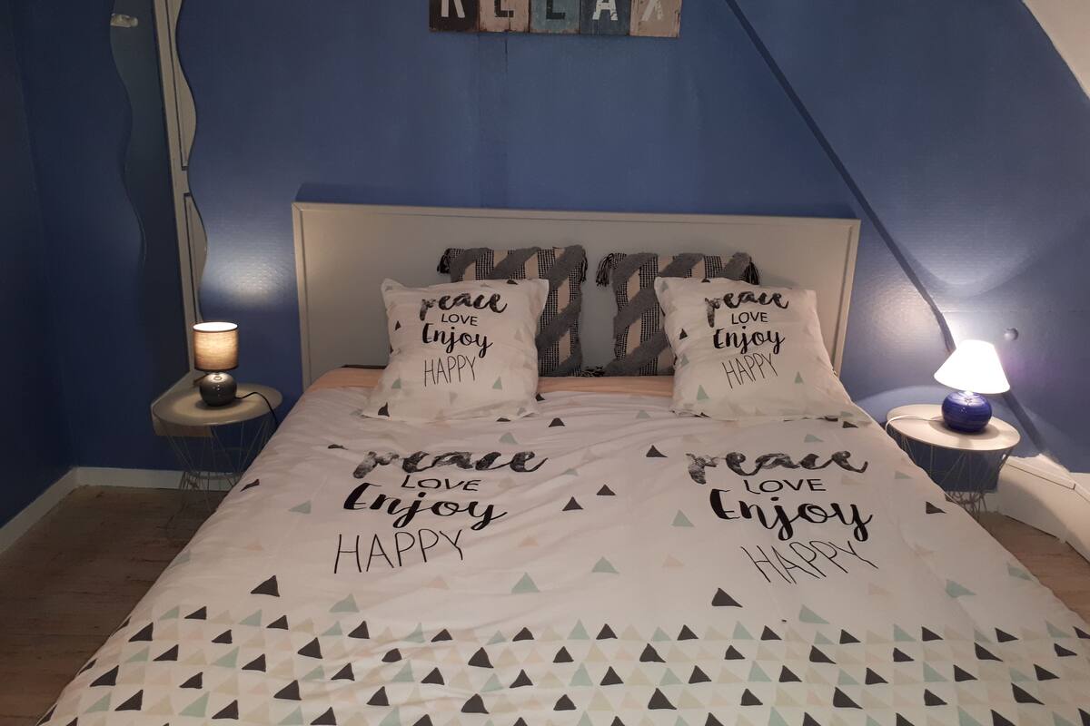 Ein Schlafzimmer mit schrägem Dach, einem Bett mit geometrischem Muster auf der Bettdecke, zwei Kissen mit motivierenden Sprüchen, einer Nachttischlampe und einem an der Wand befestigten Schild mit der Aufschrift 'RELAX'.