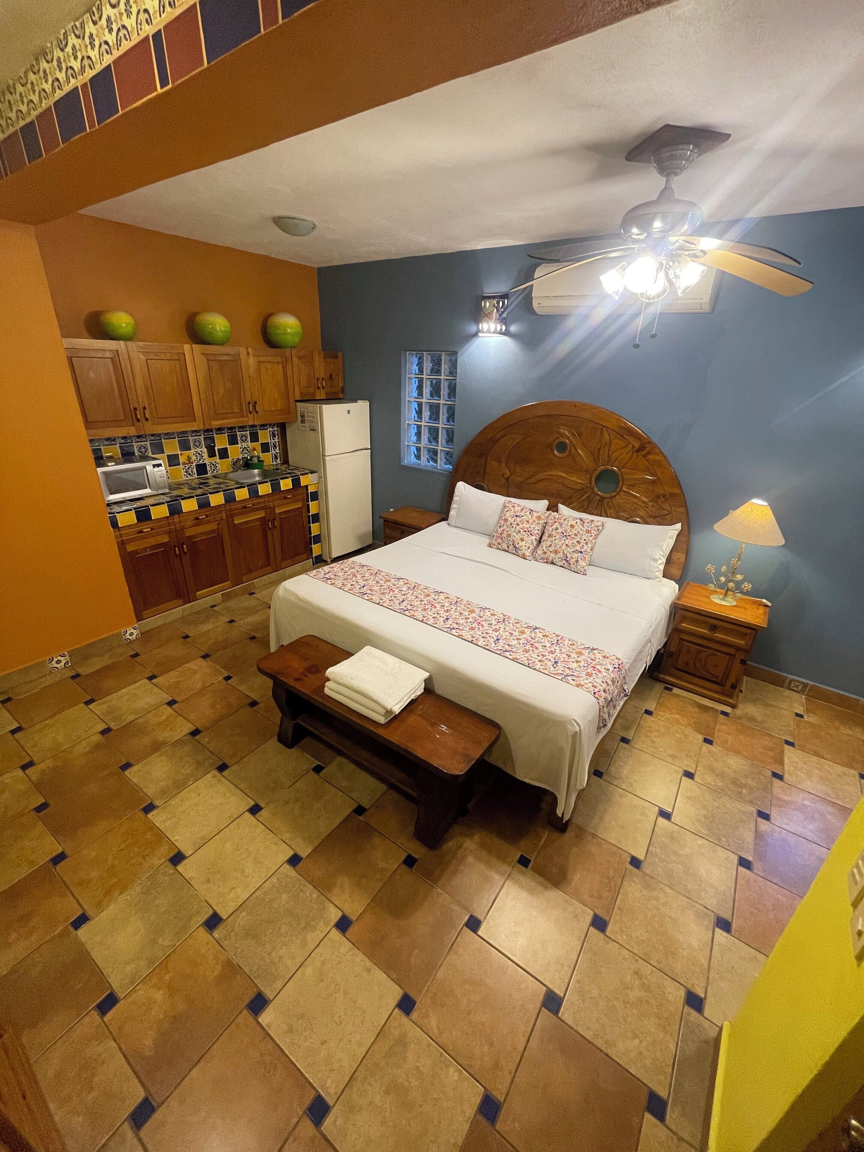 A bedroom with a bed, a wooden headboard, a nightstand, a bench, a ceiling fan, a window, and a kitchen area with cabinets and a refrigerator.
