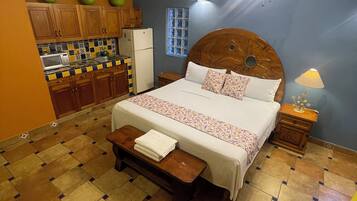 A bedroom with a bed, a wooden headboard, a nightstand, a bench, a ceiling fan, a window, and a kitchen area with cabinets and a refrigerator.