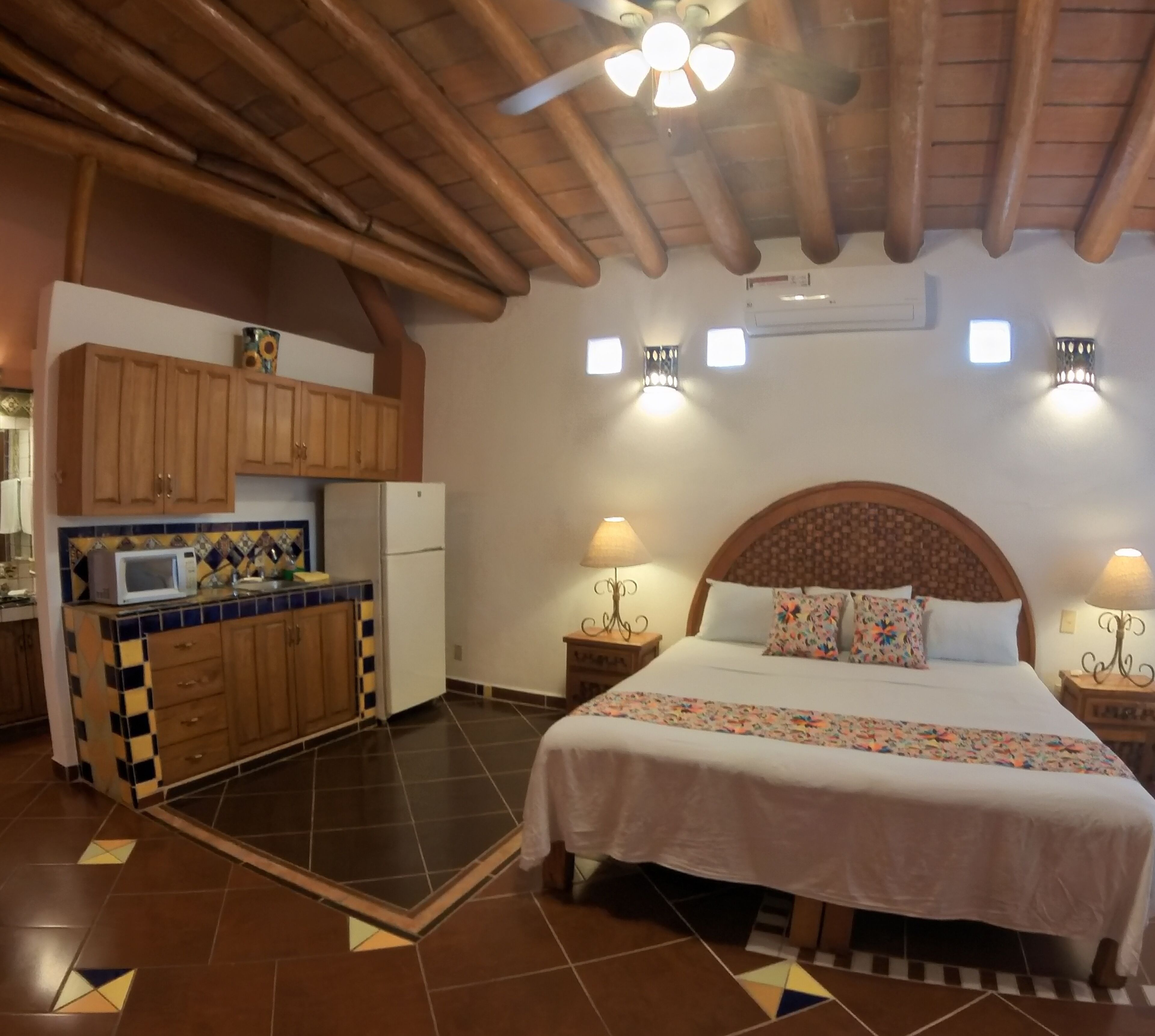 Un dormitorio con una cama grande, techo de madera, zona de cocina con armarios y un refrigerador, y dos lámparas de noche.