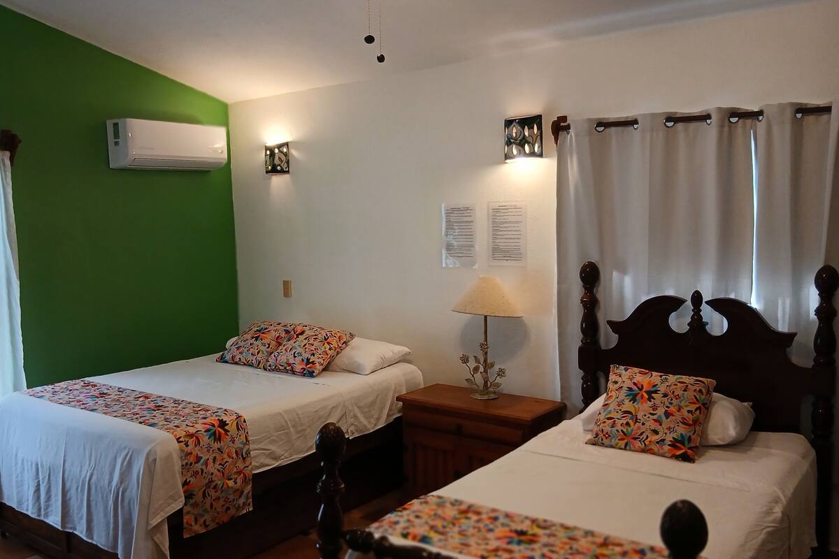 A room with two beds, a green wall, an air conditioner, a nightstand, a lamp, and a window with curtains.
