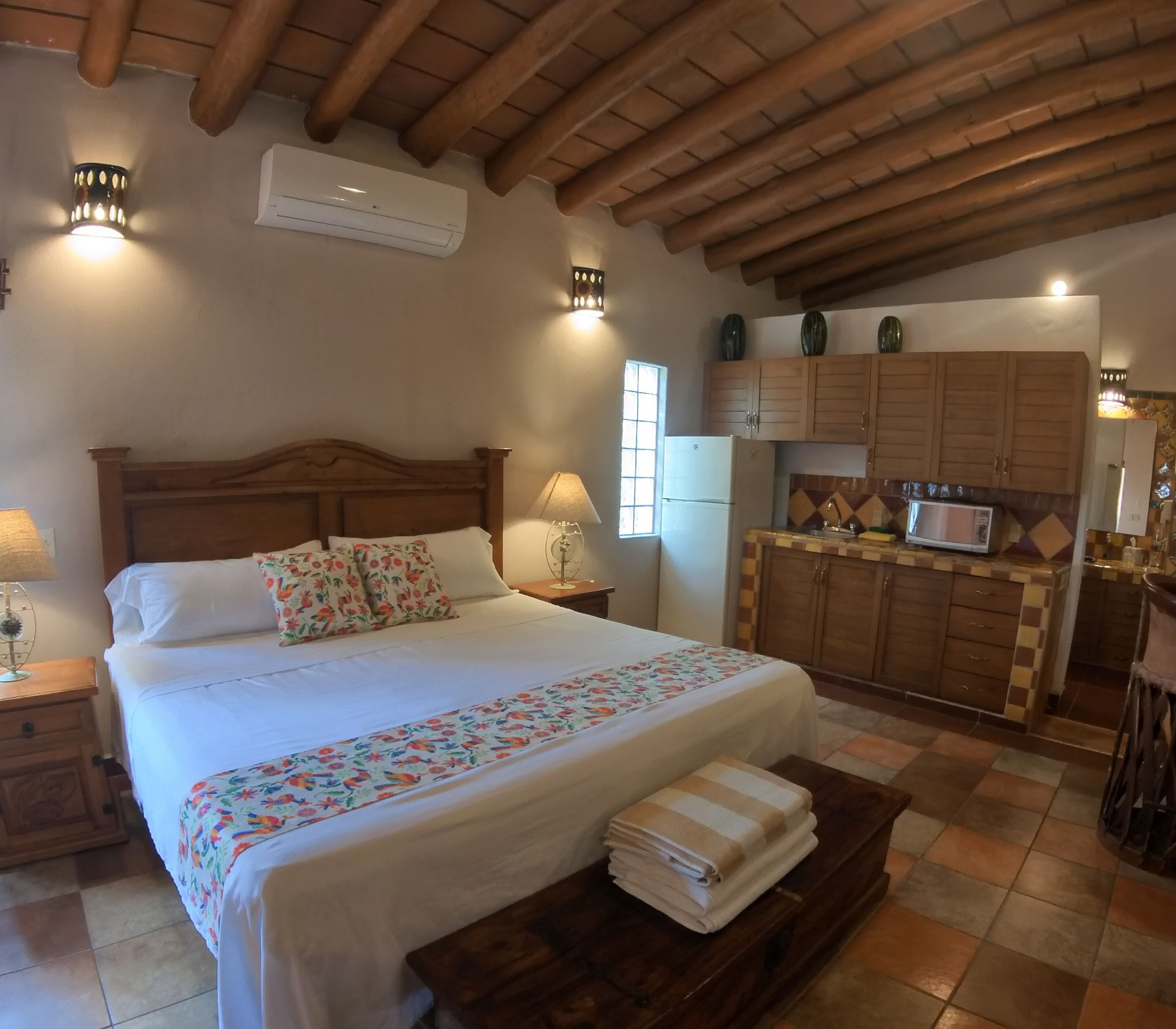 Un dormitorio con cabecera de madera, cama con ropa de cama floral, una lámpara de noche, una mesita de noche, una pequeña mesa con toallas y una zona de cocina con armarios de madera y microondas.