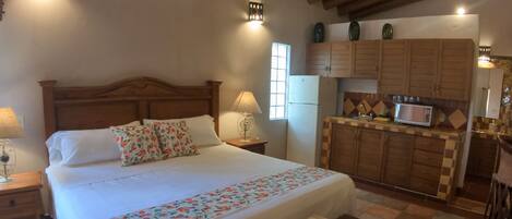 Un dormitorio con cabecera de madera, cama con ropa de cama floral, una lámpara de noche, una mesita de noche, una pequeña mesa con toallas y una zona de cocina con armarios de madera y microondas.