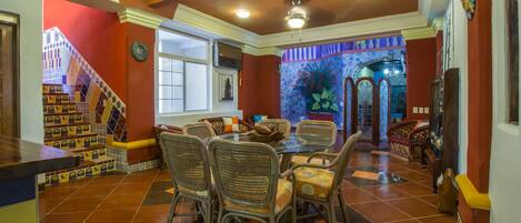 A dining area with a glass table and wicker chairs, a staircase with colorful tiles, and a view of a garden through a window.
