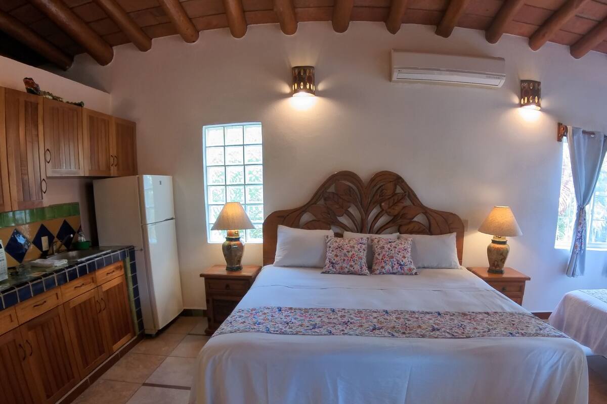 A bedroom with a wooden headboard, two bedside lamps, a bed with floral bedding, a window with curtains, and a kitchen area with wooden cabinets and a refrigerator.