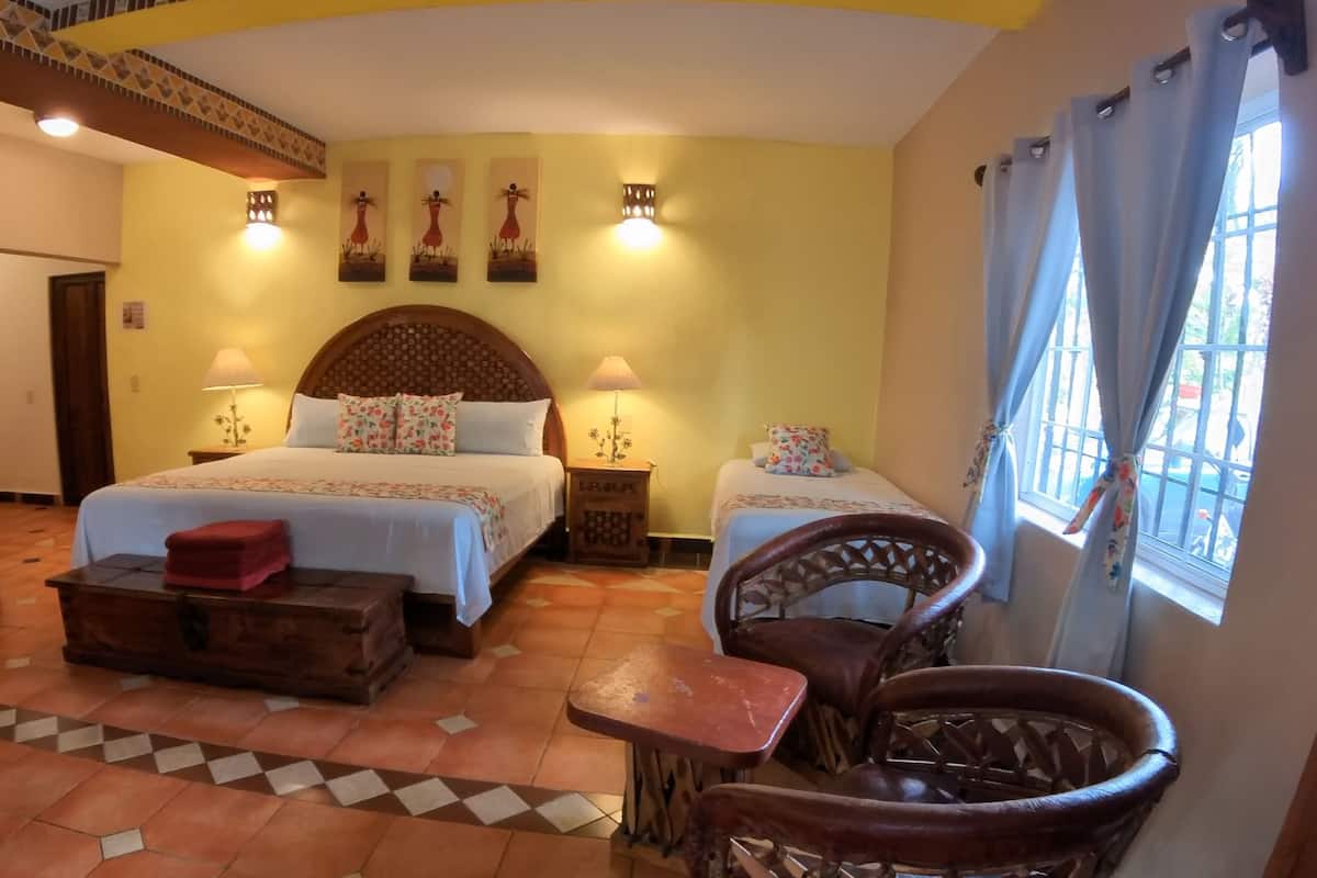 A hotel room with two beds, a dining table, wicker chairs, and a view of the outside.
