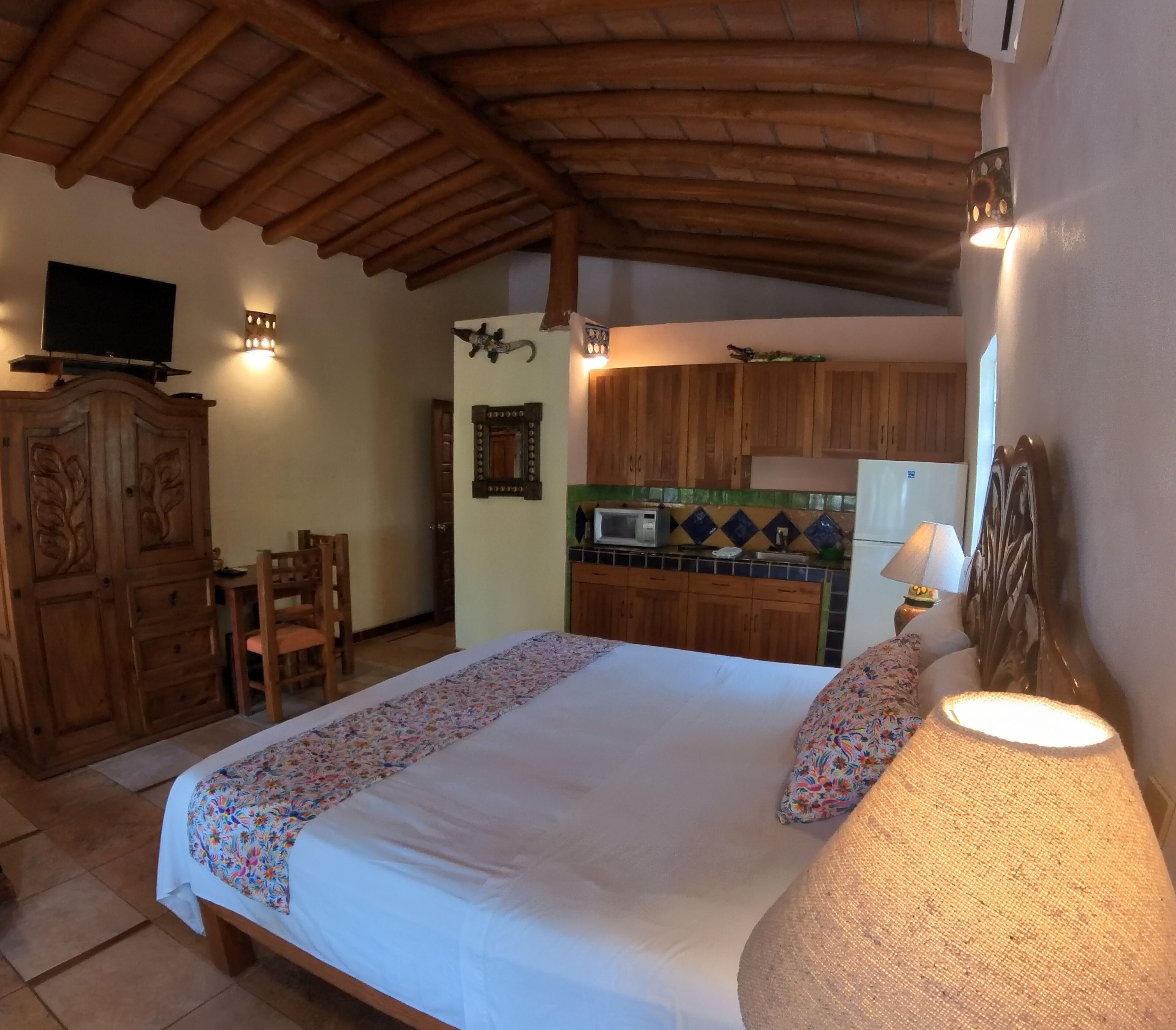 Une chambre avec un grand lit, une armoire en bois, une télévision et un coin cuisine équipé de placards et d’un four à micro-ondes.