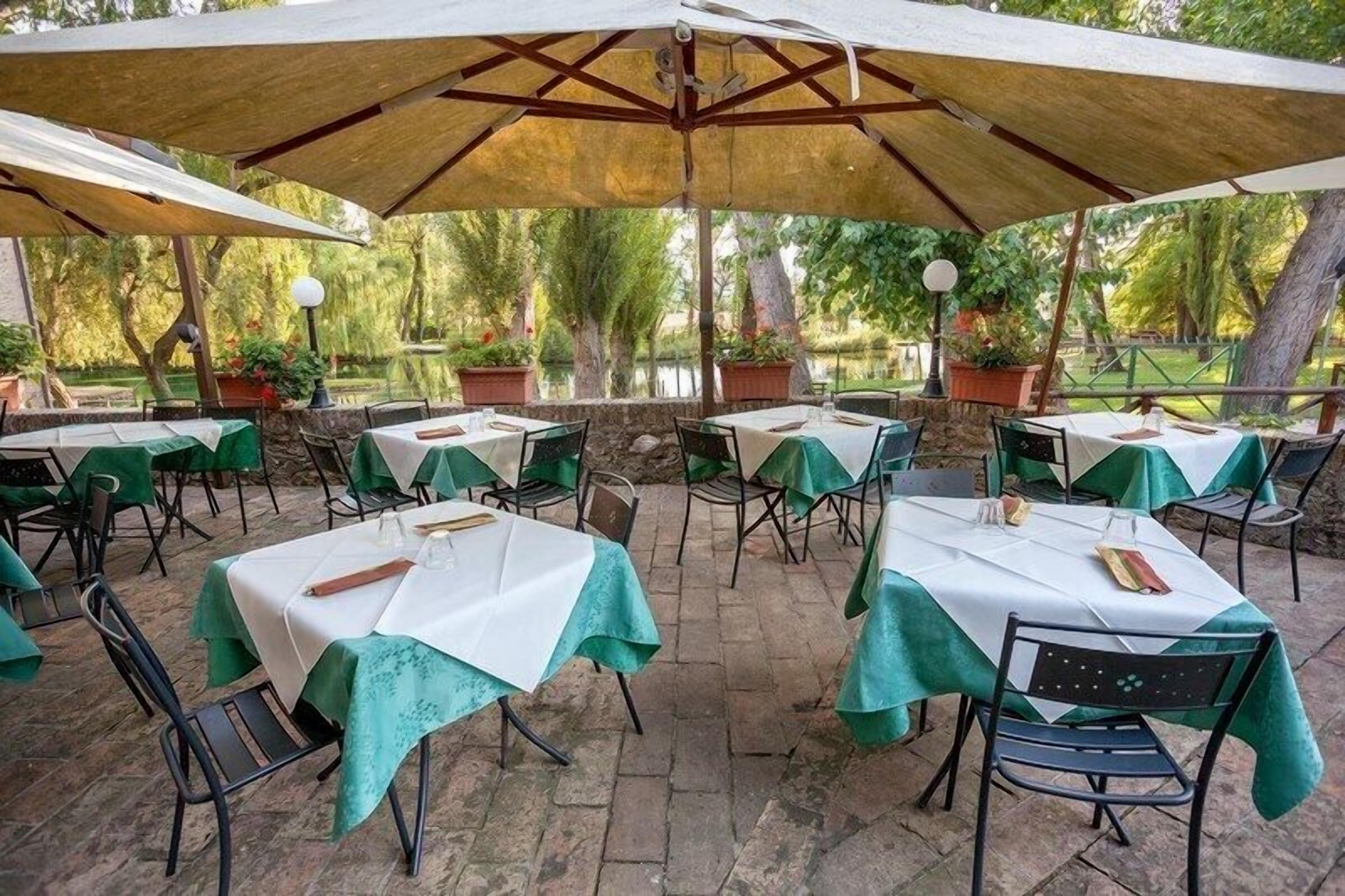 An outdoor dining area with tables covered in white tablecloths and green chair covers, surrounded by stone walls and potted plants.