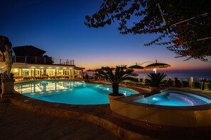 A swimming pool area with a hot tub, outdoor seating, and a view of the ocean at dusk.
