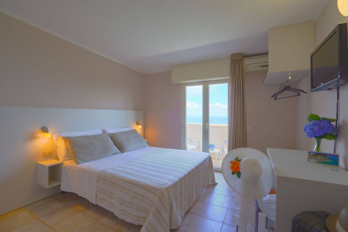 A hotel room with a bed, bedside table, lamp, desk, chair, and a view of the sea through the window.