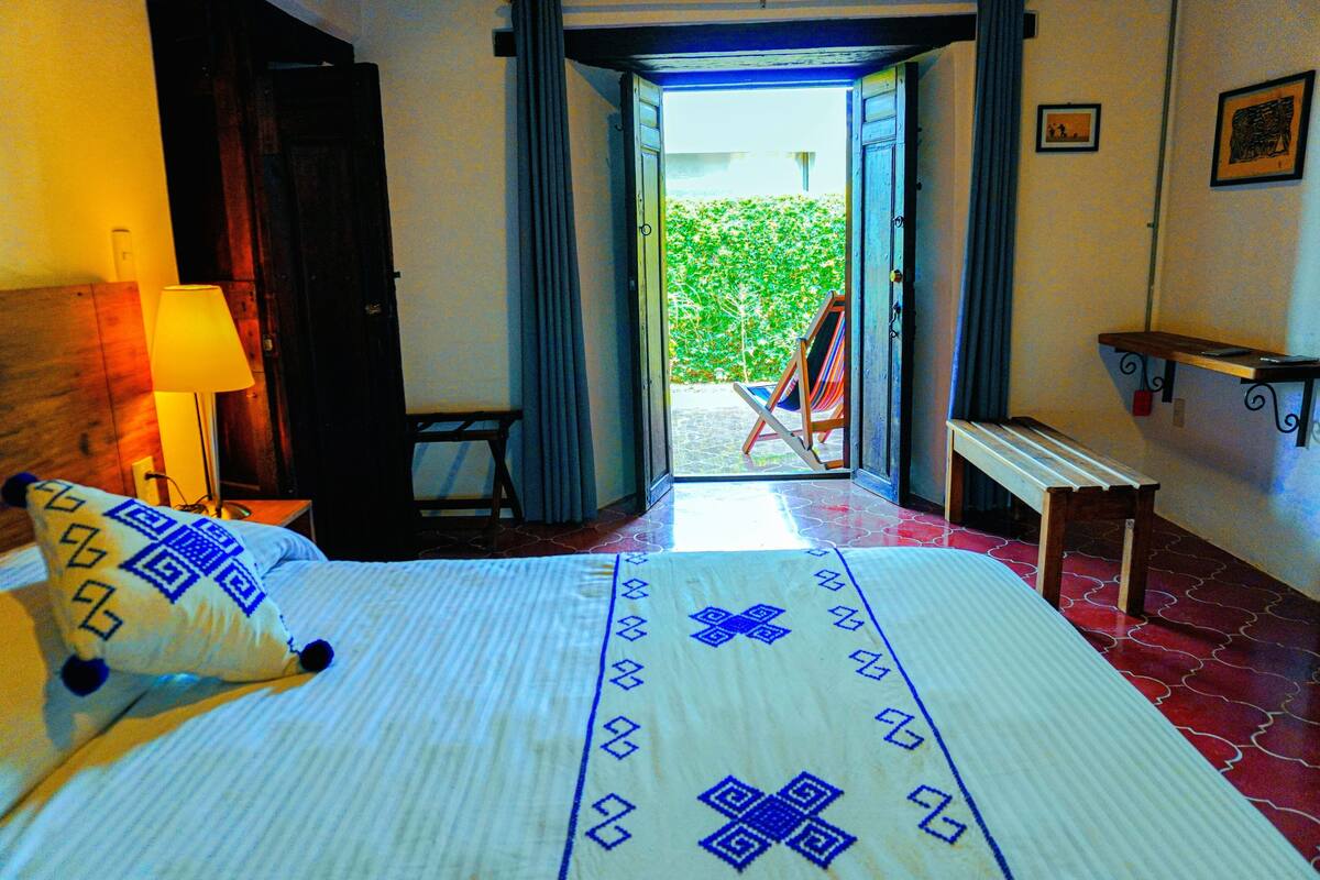 Una habitación de hotel con una cama, una lámpara, un banco y una puerta abierta a un jardín.
