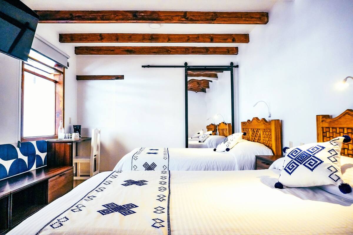 Habitación de hotel con dos camas, vigas de madera en el techo, una mesita con una silla y una ventana con persianas.