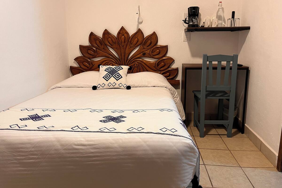 Una cama pequeña y bien hecha con un cabecero decorativo, una silla y un estante con artículos de aseo.