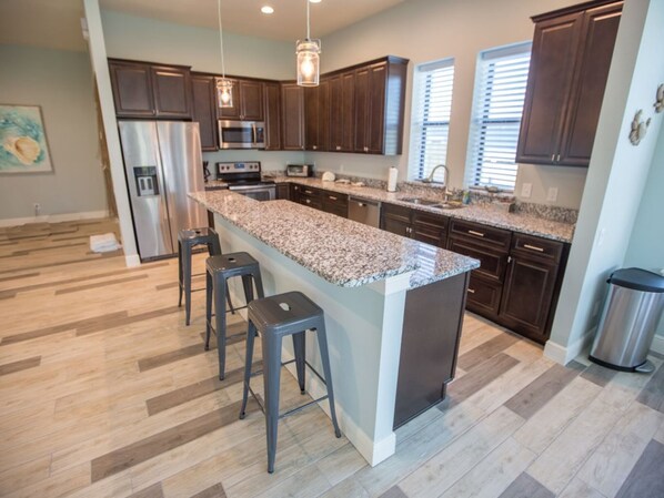 A modern kitchen with dark wood cabinets, a granite countertop island, stainless steel appliances, and bar stools. - Luna: pool, 1 blk to the beach. 3b/3.5ba. beach items, longbrd, picklball close (Cocoa Beach)