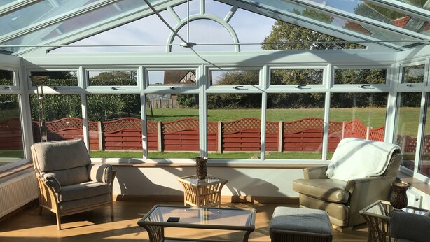 Une véranda dotée d’un toit en verre, aménagée avec des canapés et une table basse, donnant sur un jardin délimité par une clôture rouge.